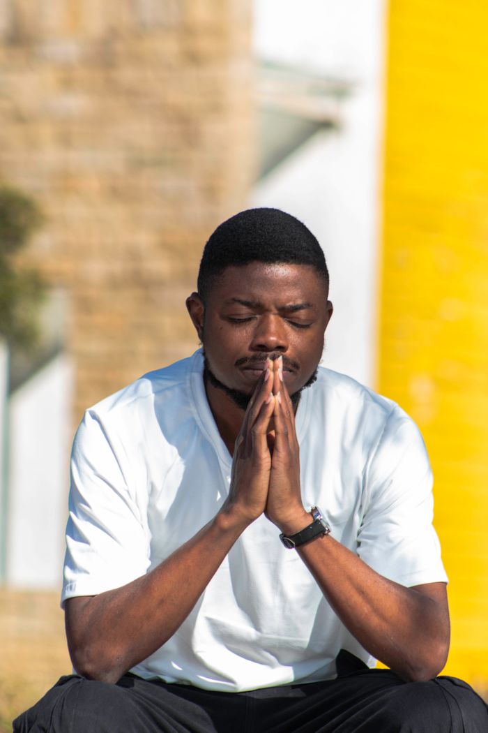 Young man sitting outdoors with eyes closed, hands clasped in prayer or meditation.